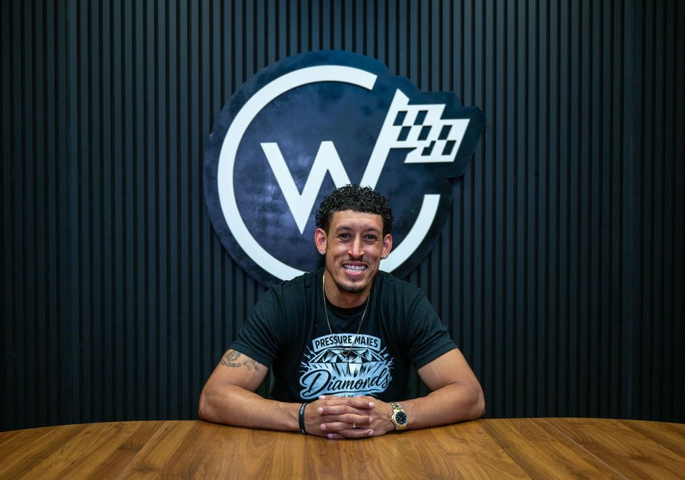 Smiling young man in a black t-shirt sits at a wooden table in front of a logo on a dark wall.