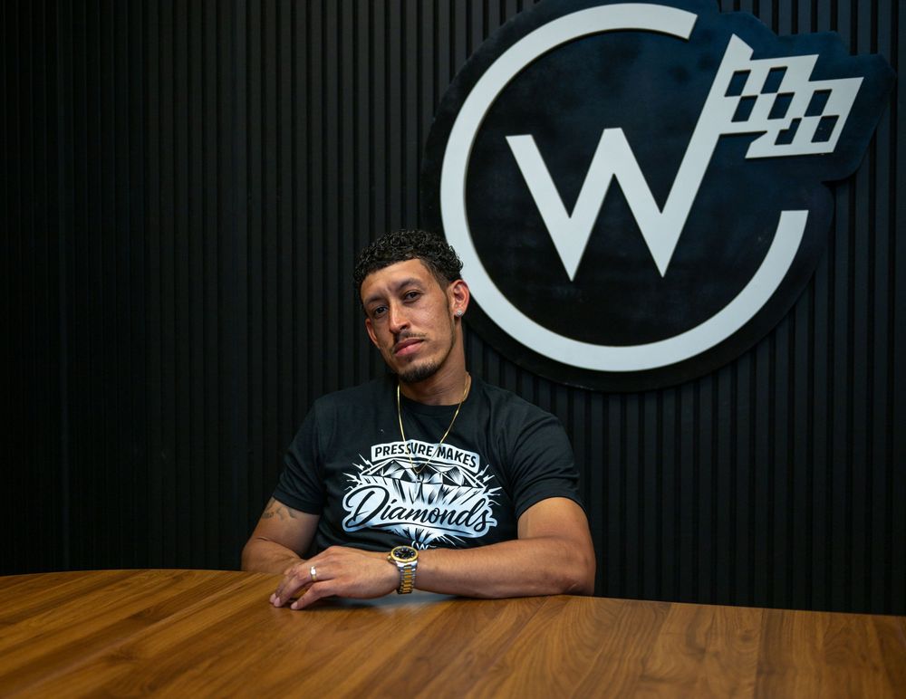 Man in black graphic tee sits at table in front of decorative W logo sign in modern interior.