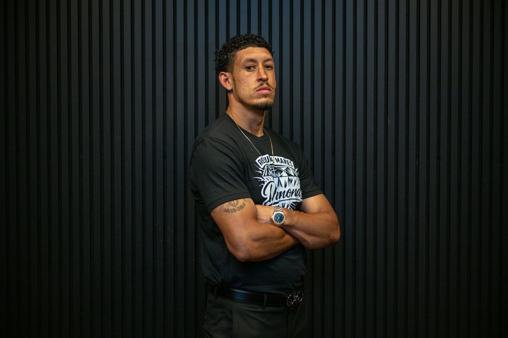 Man in black graphic t-shirt stands with arms crossed against dark striped wall, wearing a blue watch.