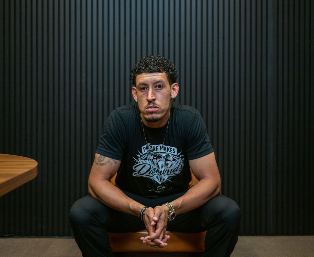 Young man in black graphic tee sits confidently against dark wood panel wall, arms resting on knees.