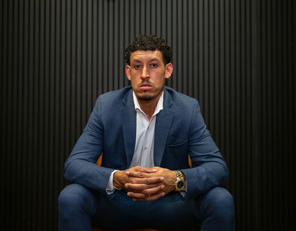 Professional man in blue suit sitting confidently in chair against dark striped background.
