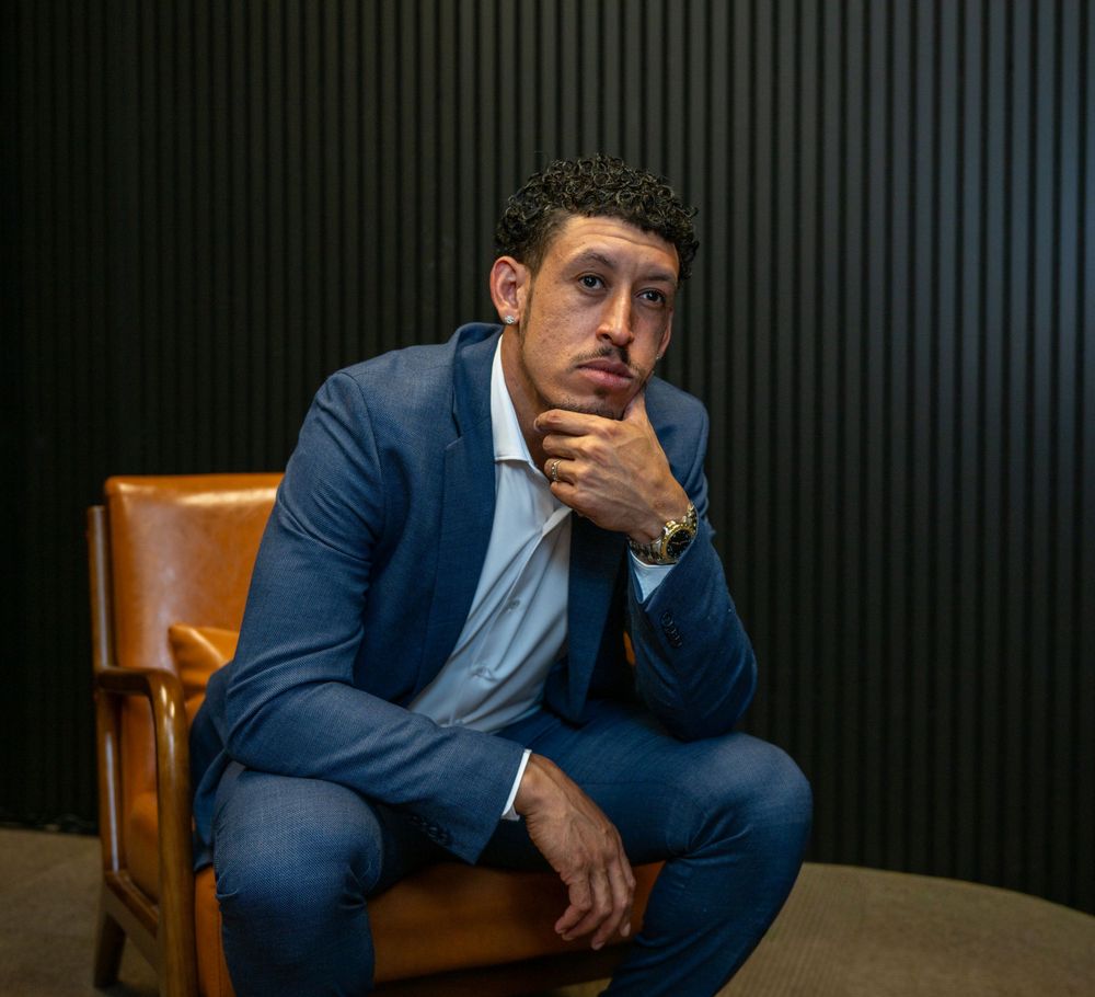 Young man in blue suit sitting in leather chair, posing thoughtfully with hand on chin in professional portrait.