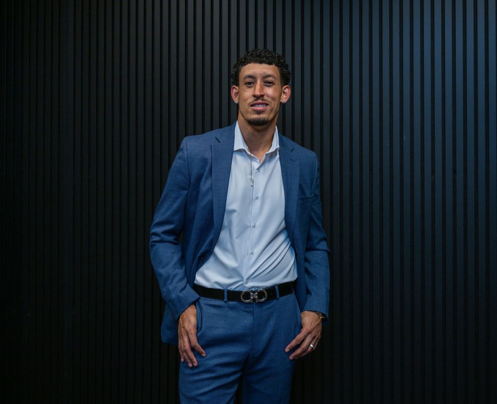 Professional man in blue suit posing confidently against dark striped wall background.