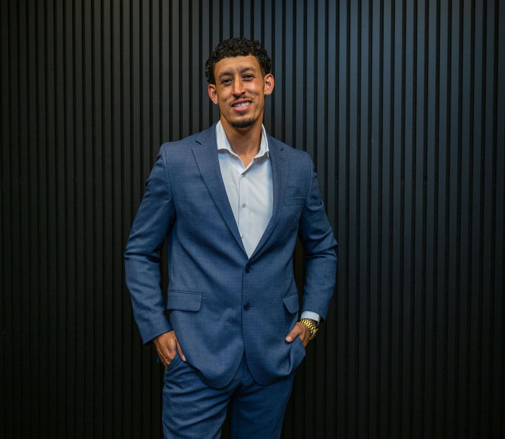 Professional man in blue suit smiling confidently against dark wall background.