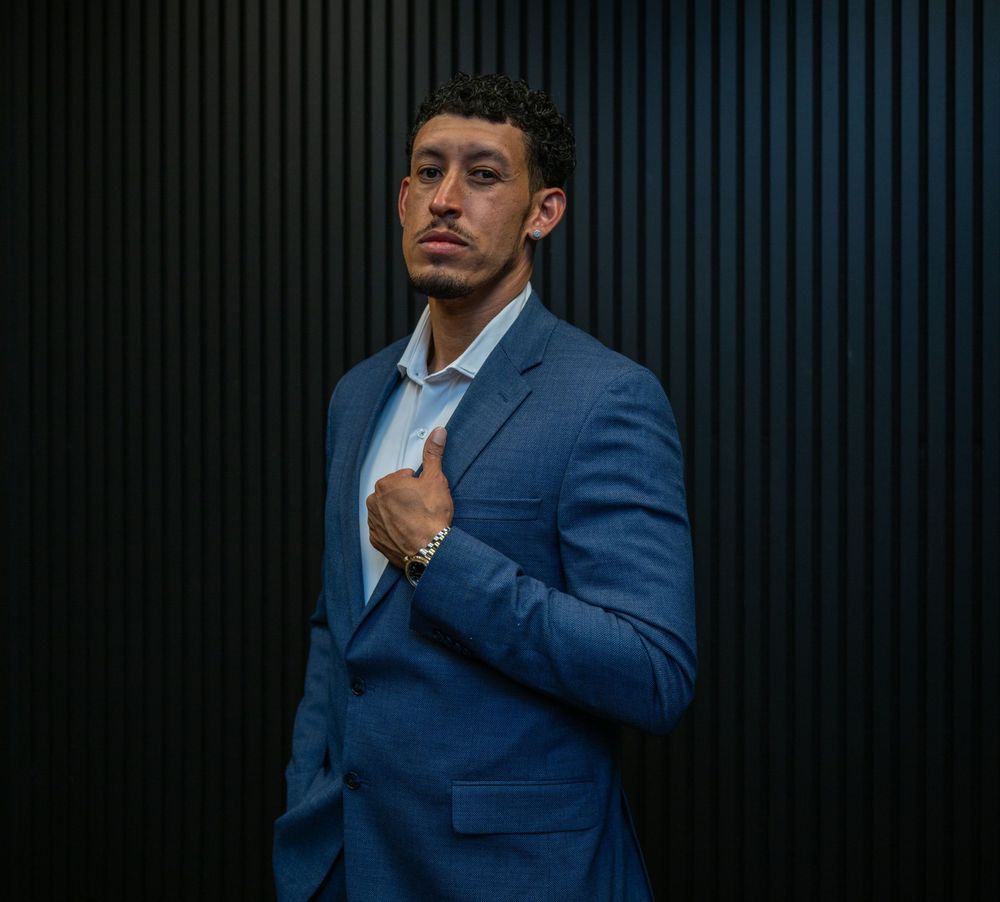 Man in blue suit posing confidently against dark striped wall, wearing a watch and light blue dress shirt.