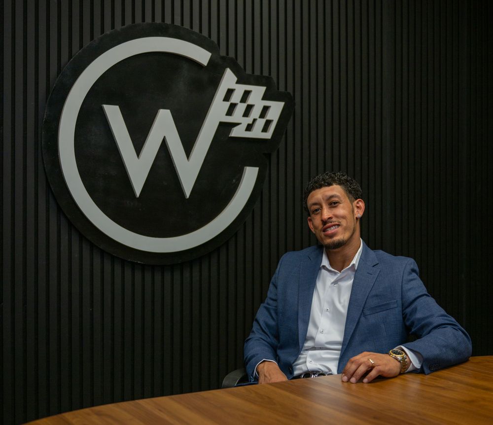 Professional man in blue suit seated at desk before a circular W logo sign in a modern office.