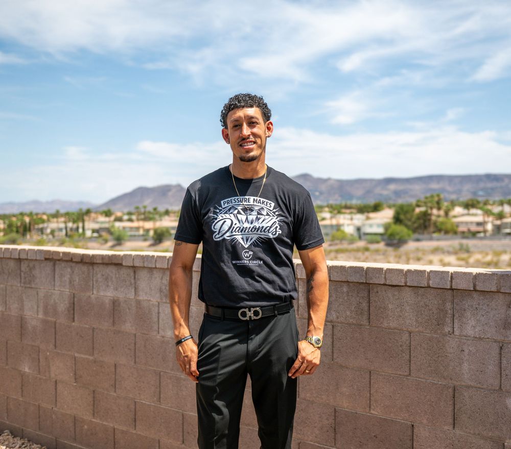 Young man in black graphic t-shirt stands confidently by a brick wall with mountain views under a blue sky.