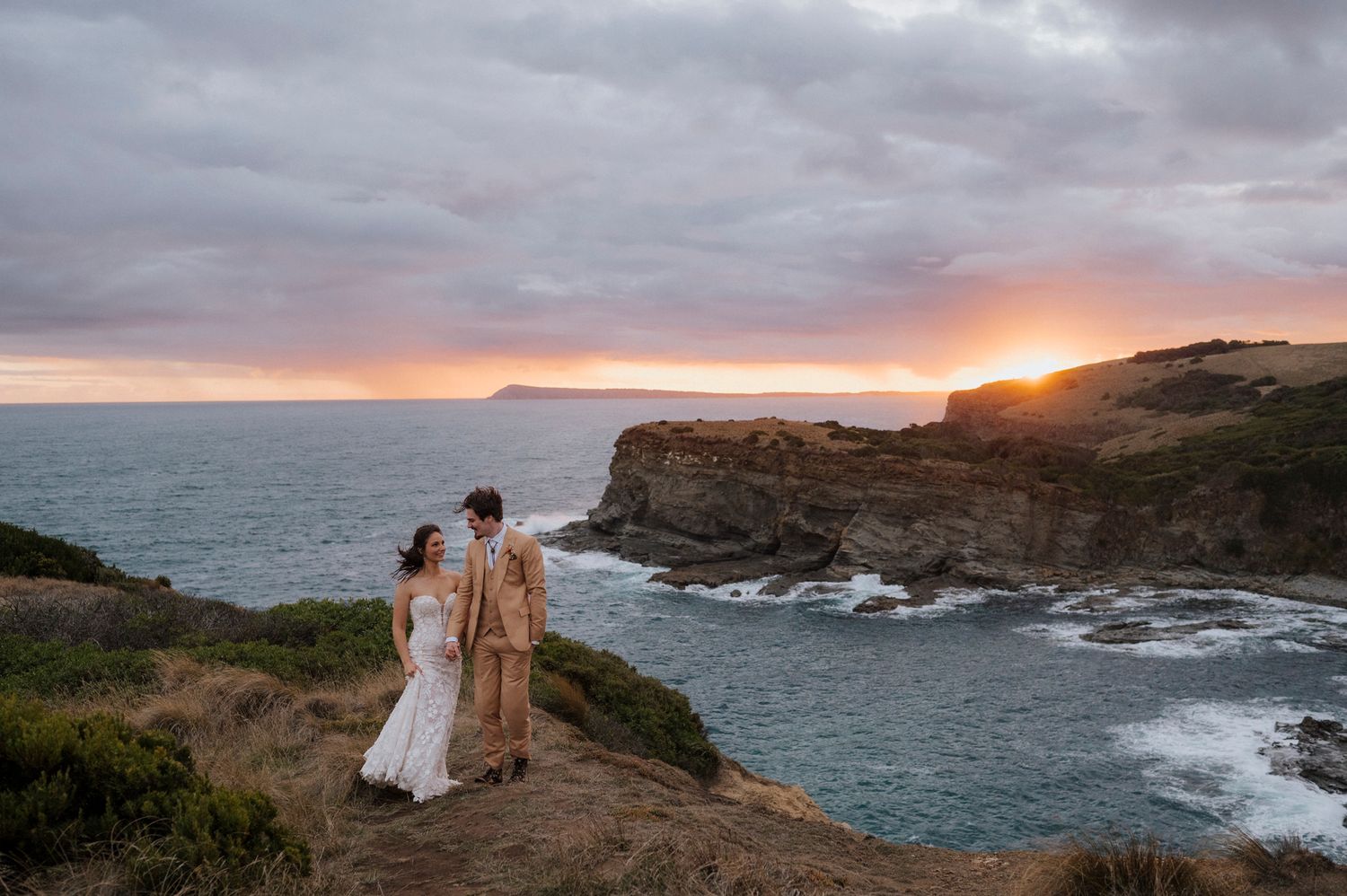 Couple in wedding attire standing on coastal cliffs at sunset with dramatic ocean and rocky headland views.