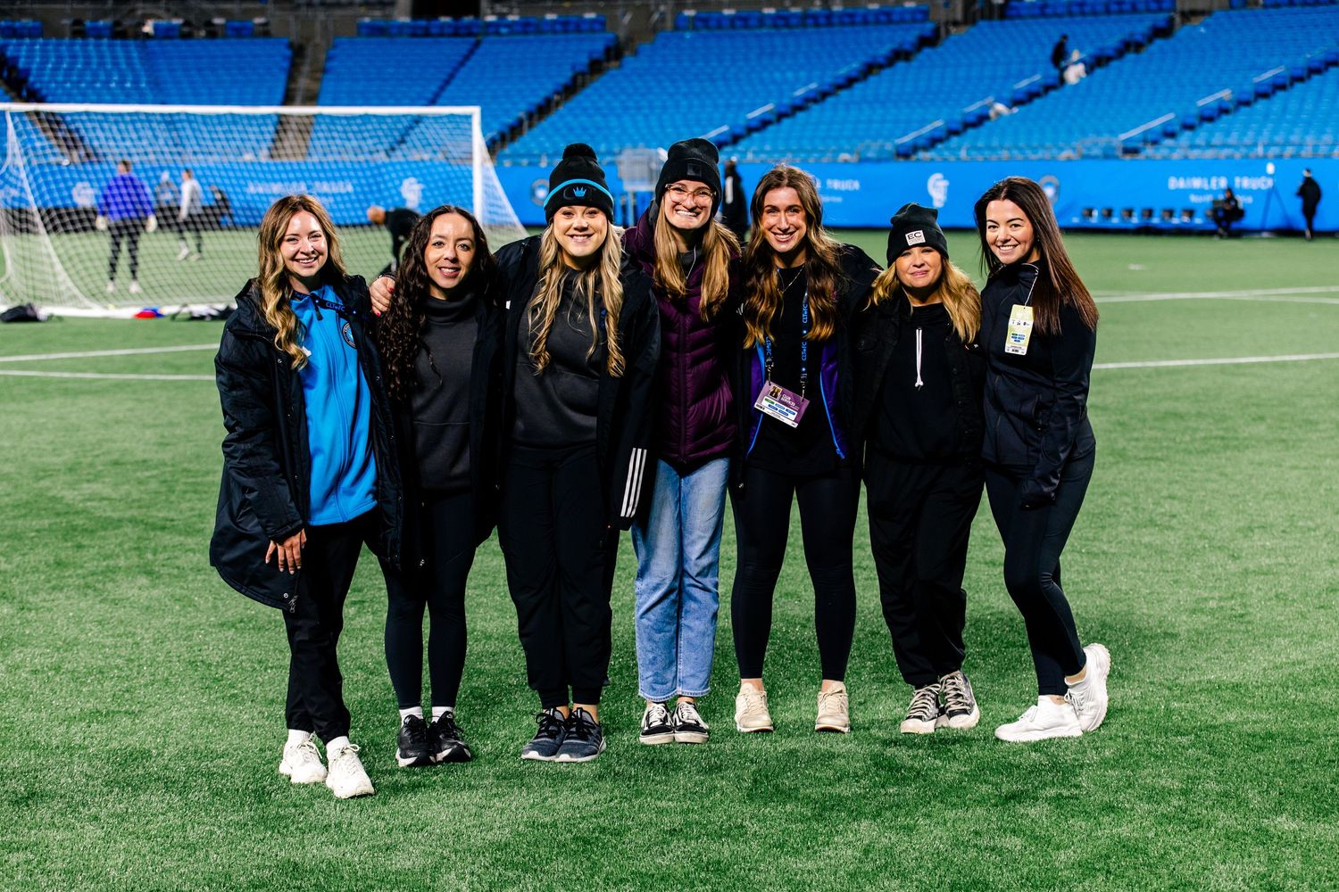 Seven women pose together smiling on a soccer field inside a large stadium at night.