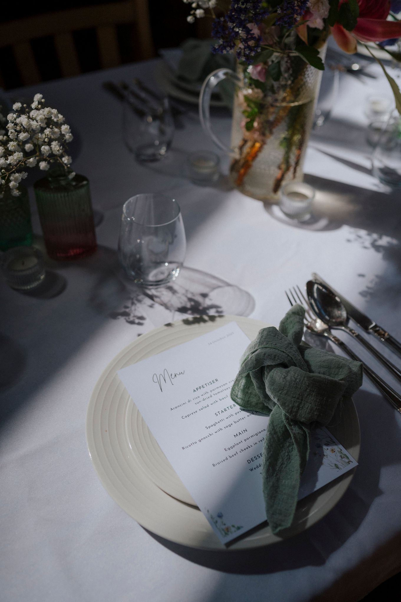 Elegant wedding table setting with green knotted napkin, menu card, silverware, and floral centerpiece on white linen.