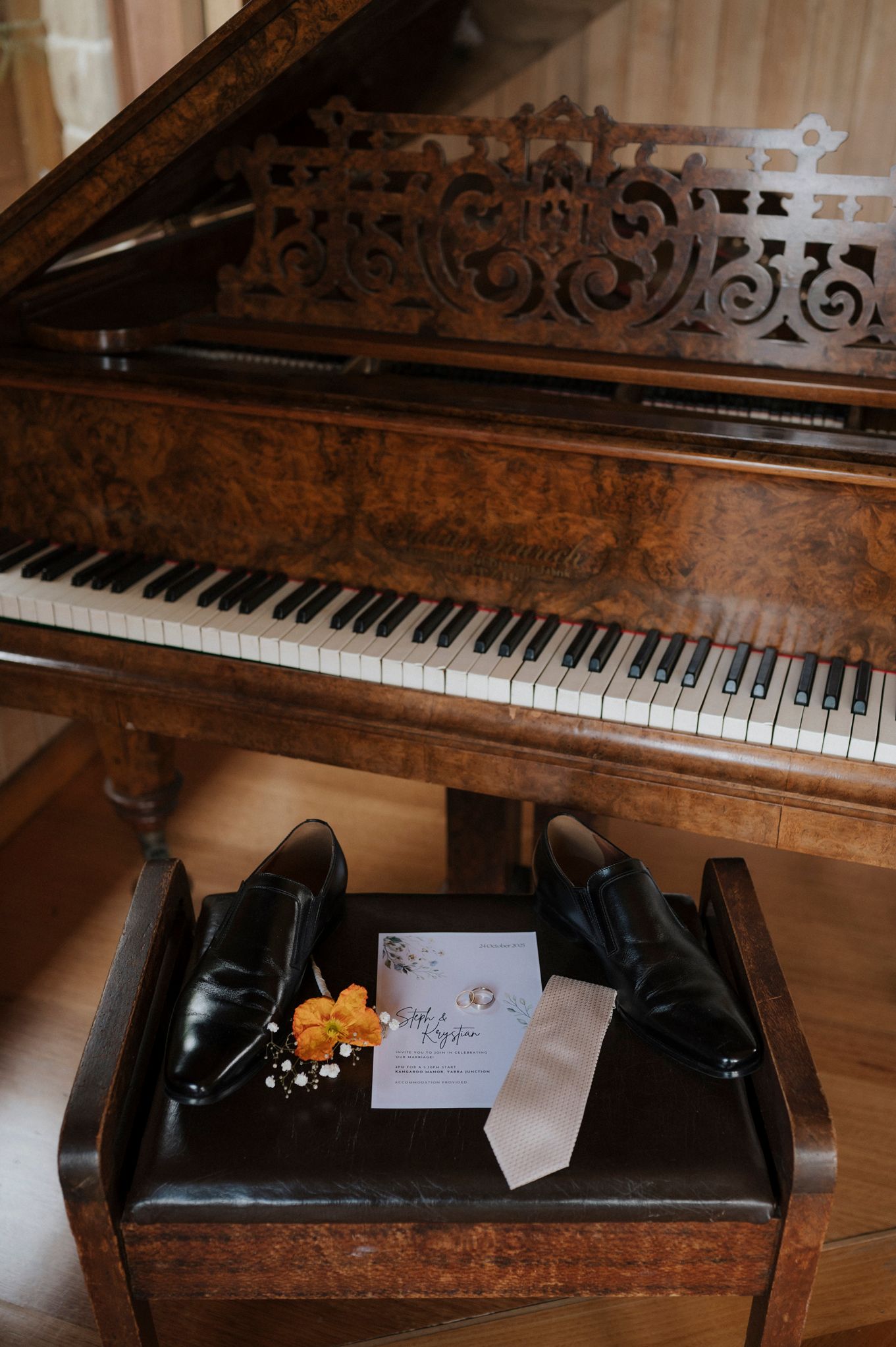Vintage grand piano with ornate woodwork, black dress shoes, boutonniere, and wedding invitation on bench.