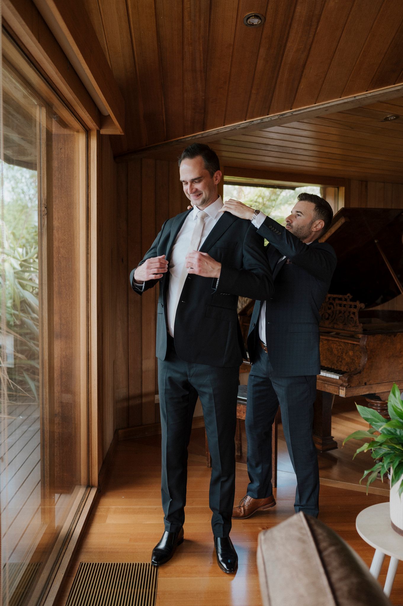 Groom being helped into dark suit jacket by groomsman in wood-paneled room with piano backdrop.