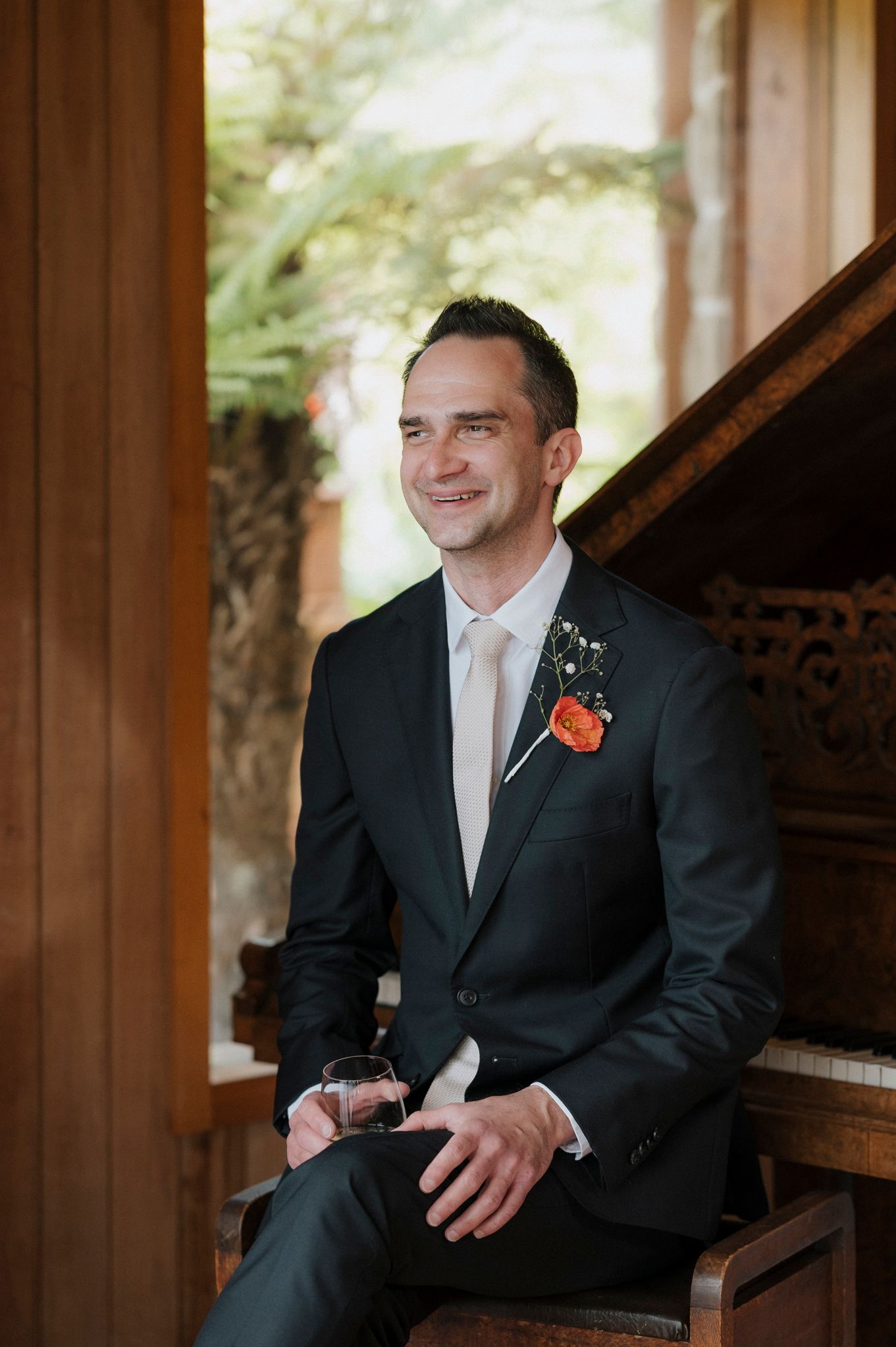Groom in dark suit with orange boutonniere sits smiling by a piano, holding a drink at a wedding venue.