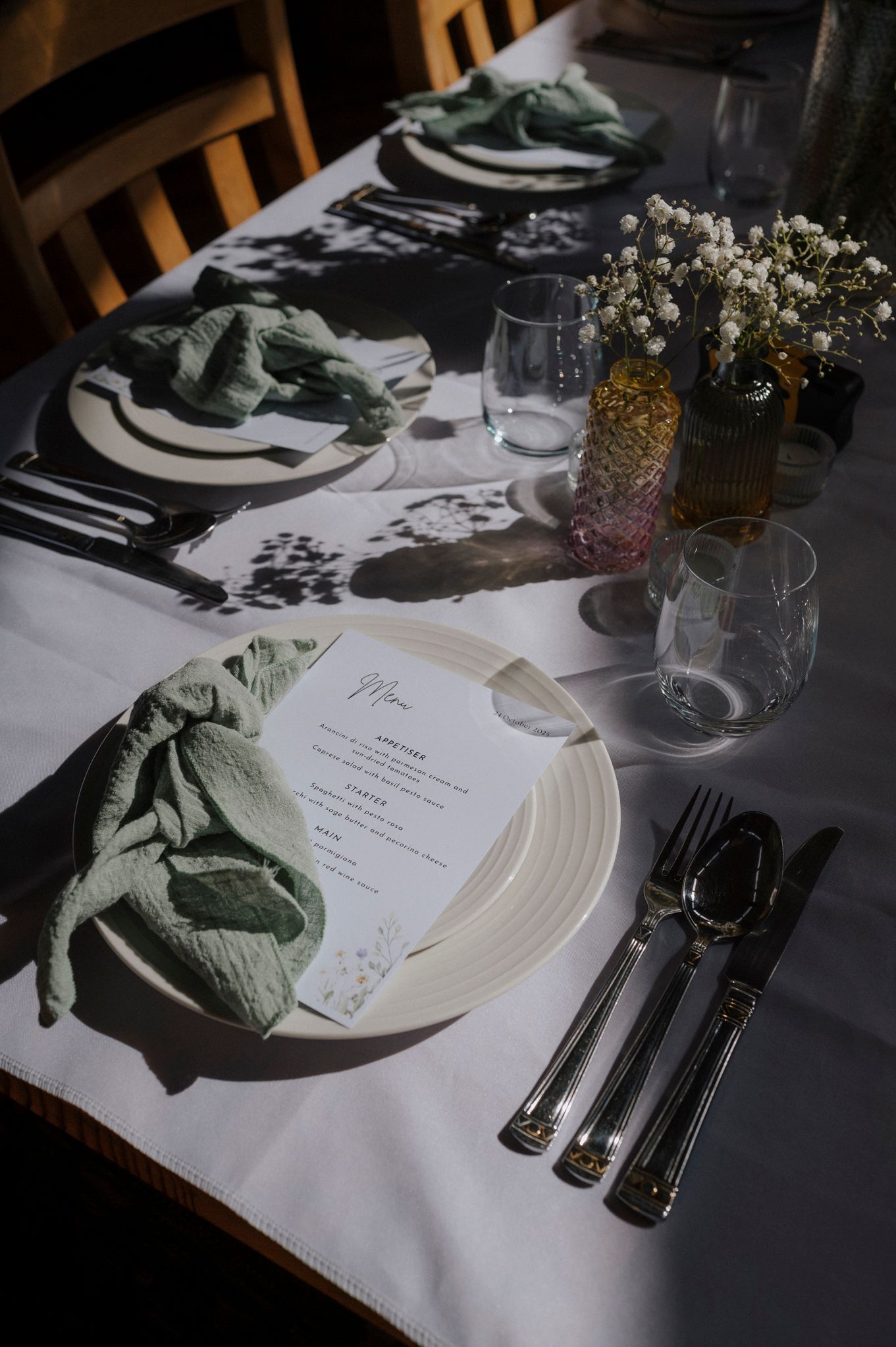 Elegant wedding table setting with sage green linen napkins, white plates, menu cards, crystal glasses, and baby's breath floral accents.