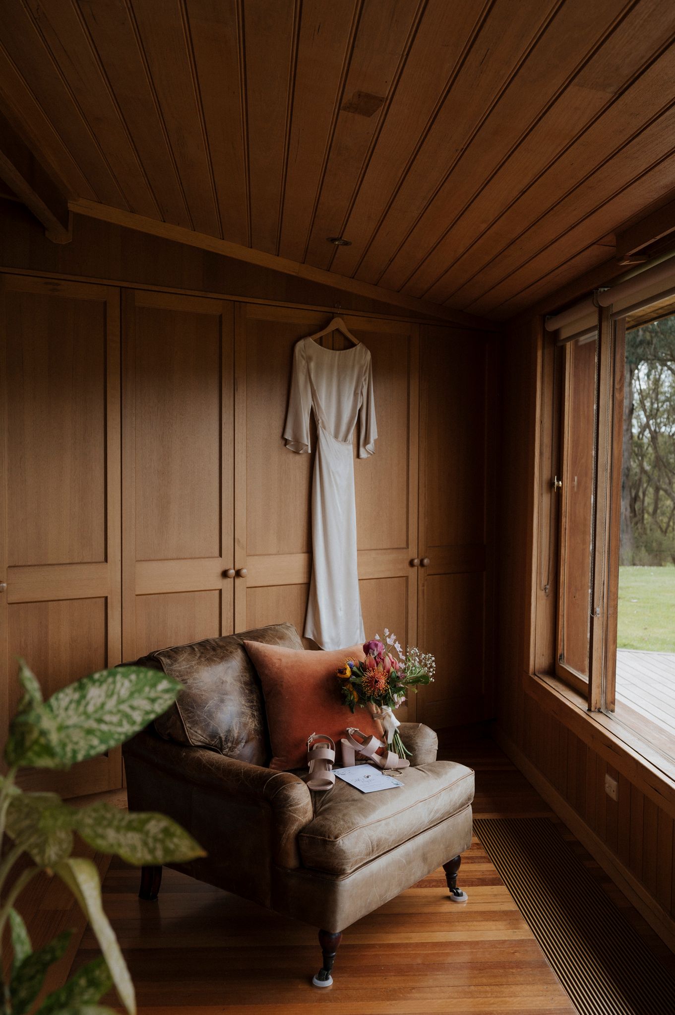 Wedding dress hanging in a warm wood-paneled room with a velvet armchair, orange pillow, and floral arrangement by a window.