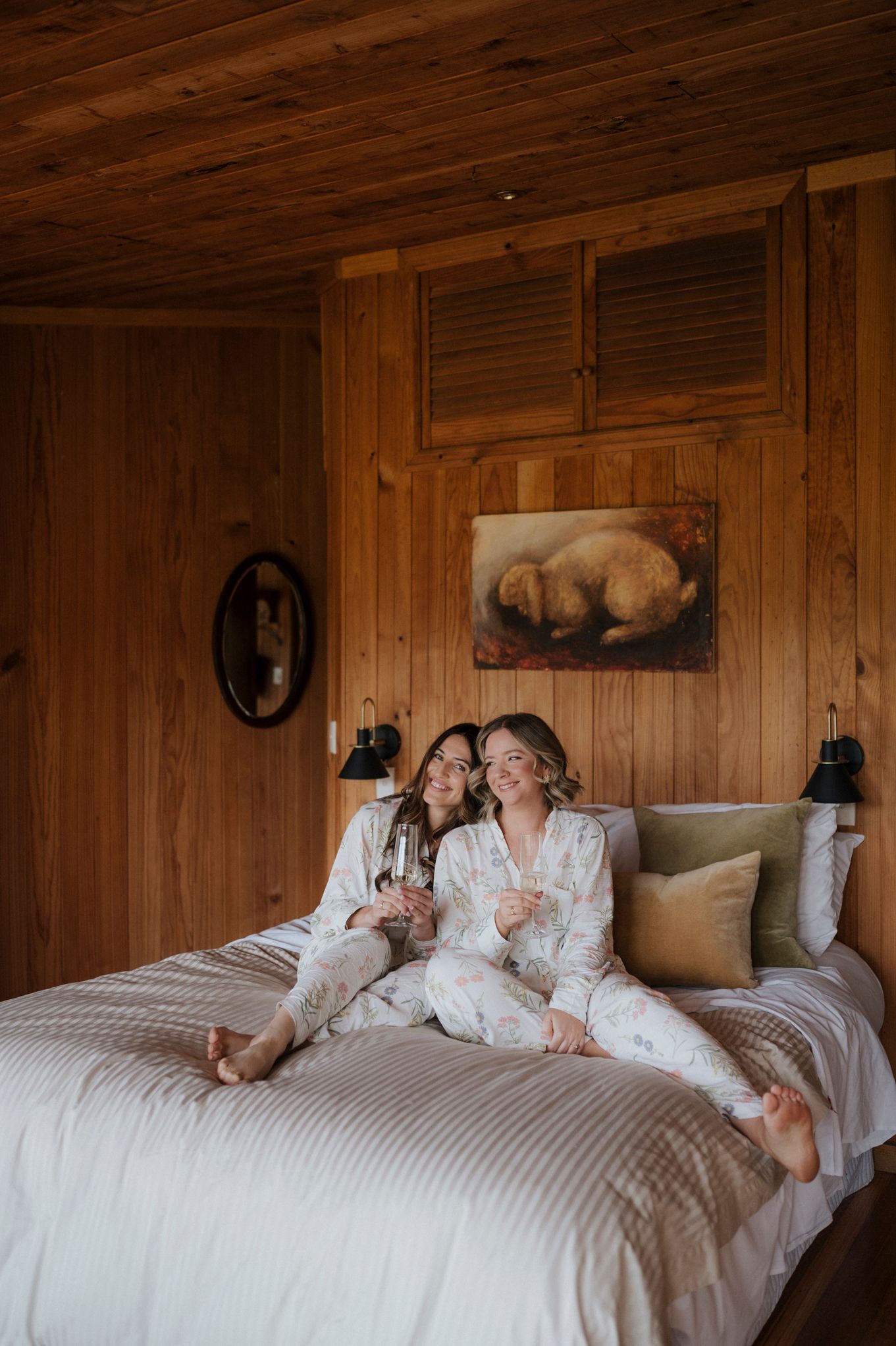 Two women in robes relaxing on a bed in a cozy wooden cabin, holding wine glasses and smiling together.