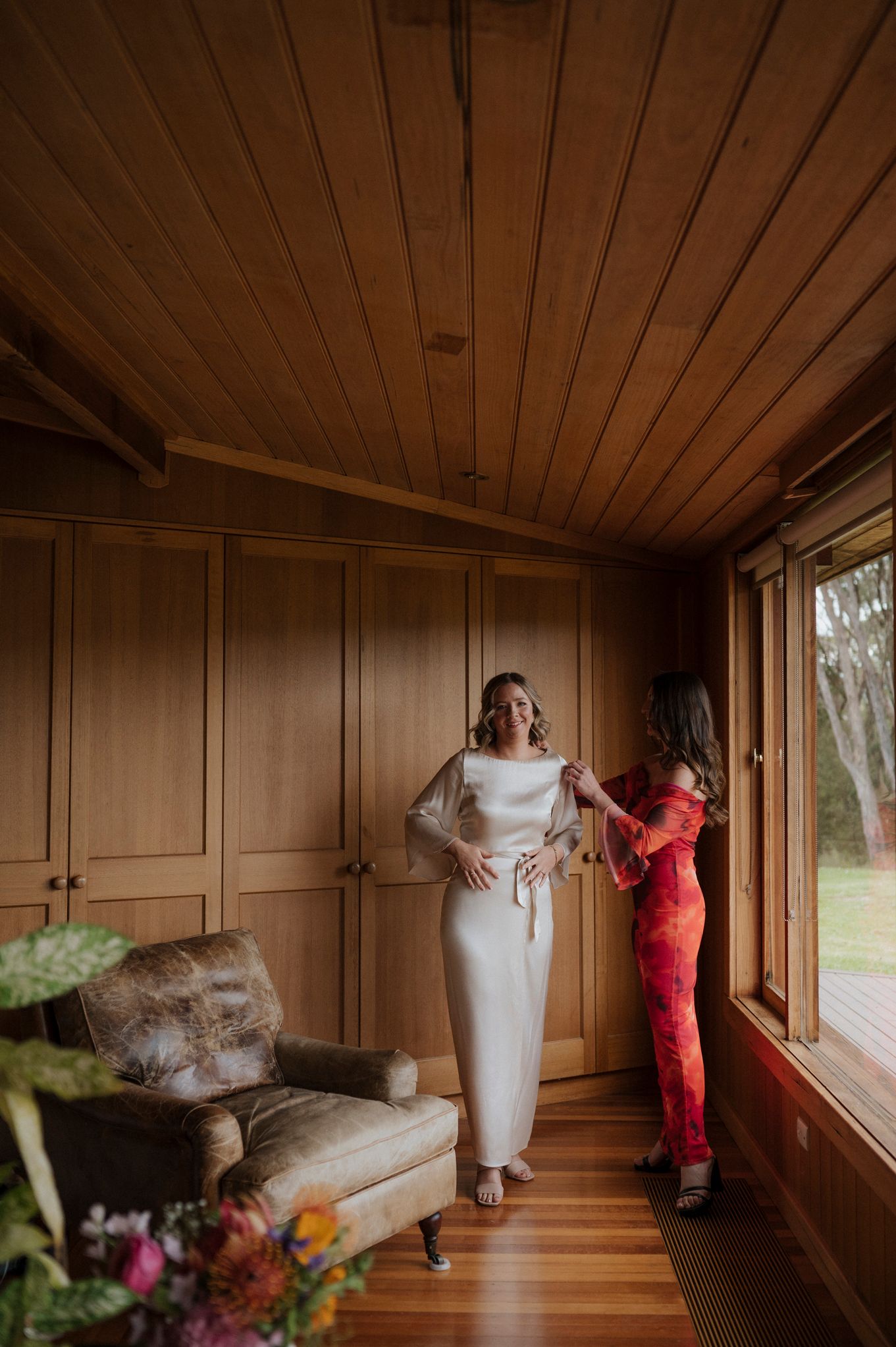 Bride in white gown getting dressed with help from bridesmaid in red floral dress in a warm wooden room.