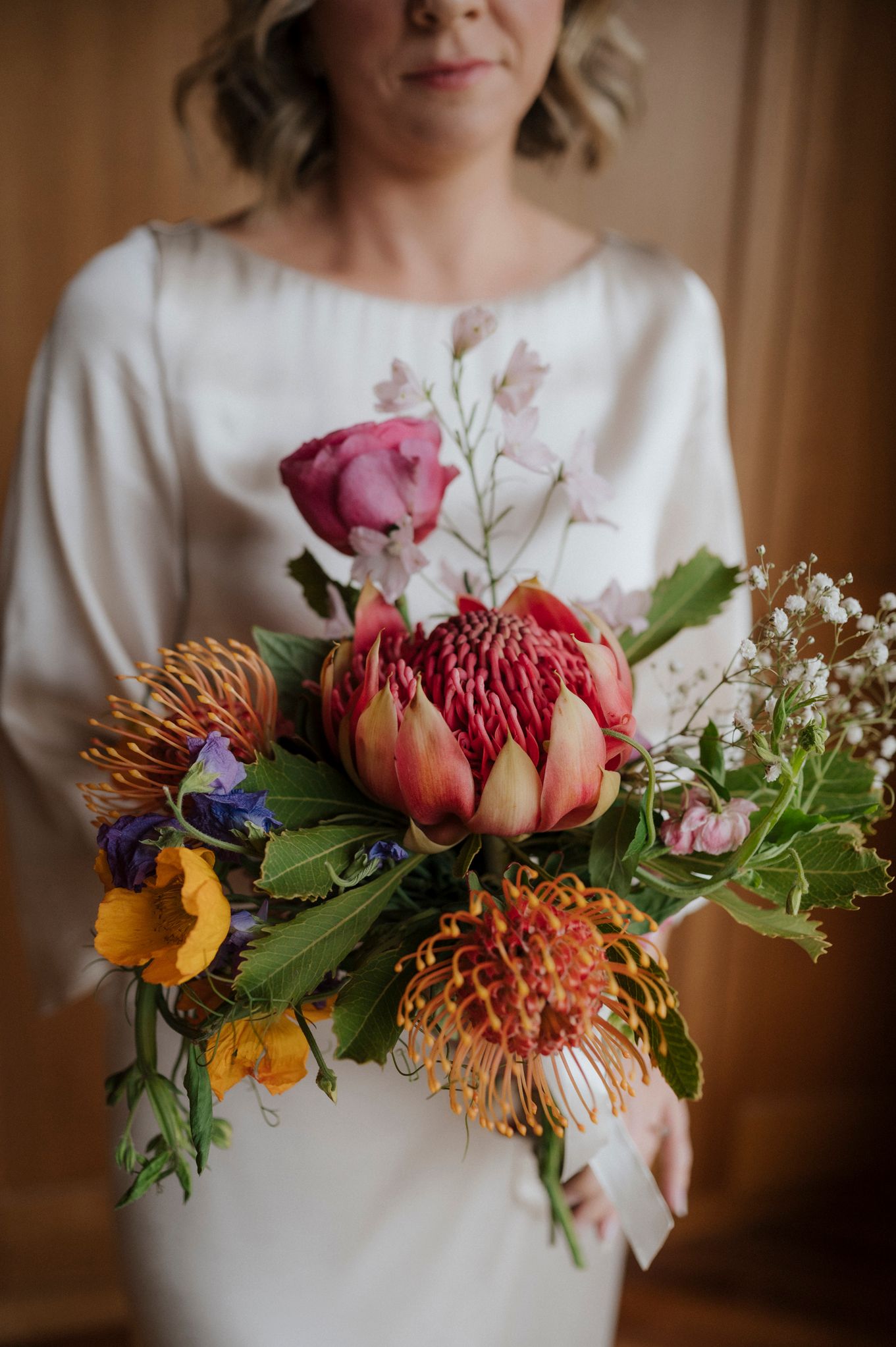 Bride holding a vibrant wedding bouquet featuring a protea, pincushion flowers, roses, and wildflowers in warm tones.