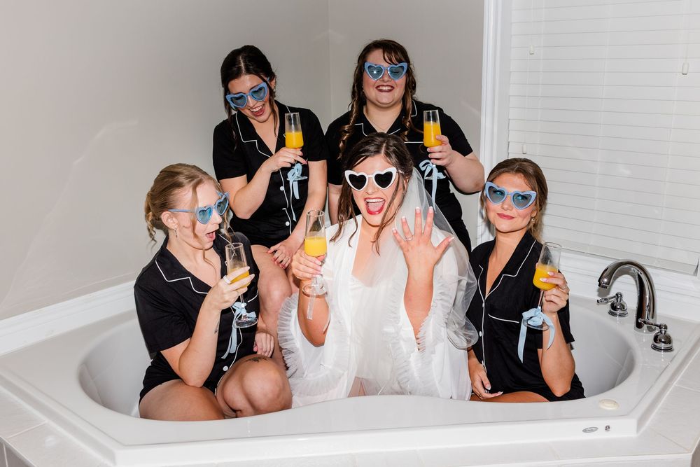 Six women in black pajamas and sunglasses celebrate a bachelorette party in a bathtub, holding champagne glasses.