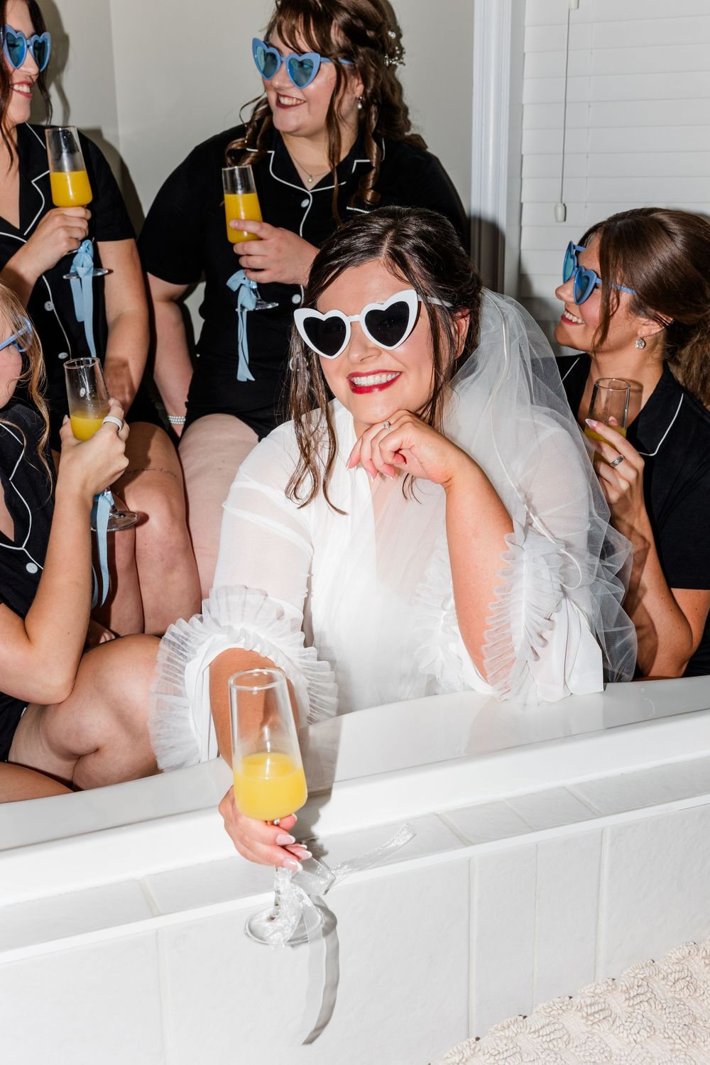 Bride in white robe, veil, and heart sunglasses holds mimosa in bathtub, surrounded by bridesmaids in black pajamas.