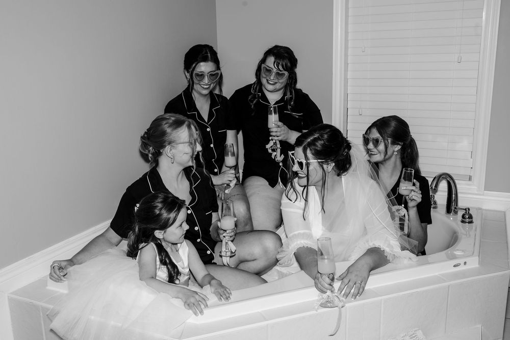 Black and white photo of bridesmaids in robes celebrating with champagne in a bathtub before a wedding.