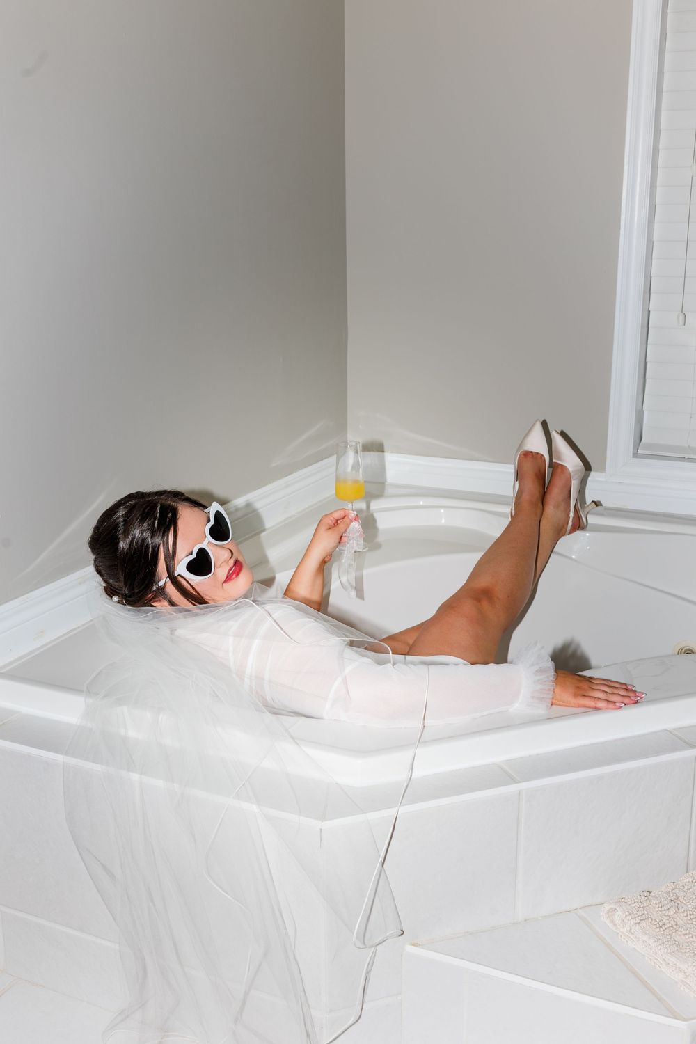 Bride in white dress and sunglasses relaxing in bathtub holding champagne glass wearing heels.