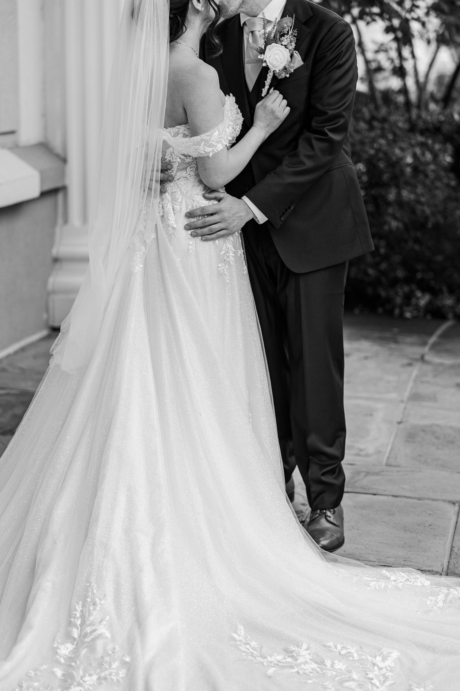 Black and white photo of a bride and groom embracing, showcasing her lace wedding gown and flowing veil.