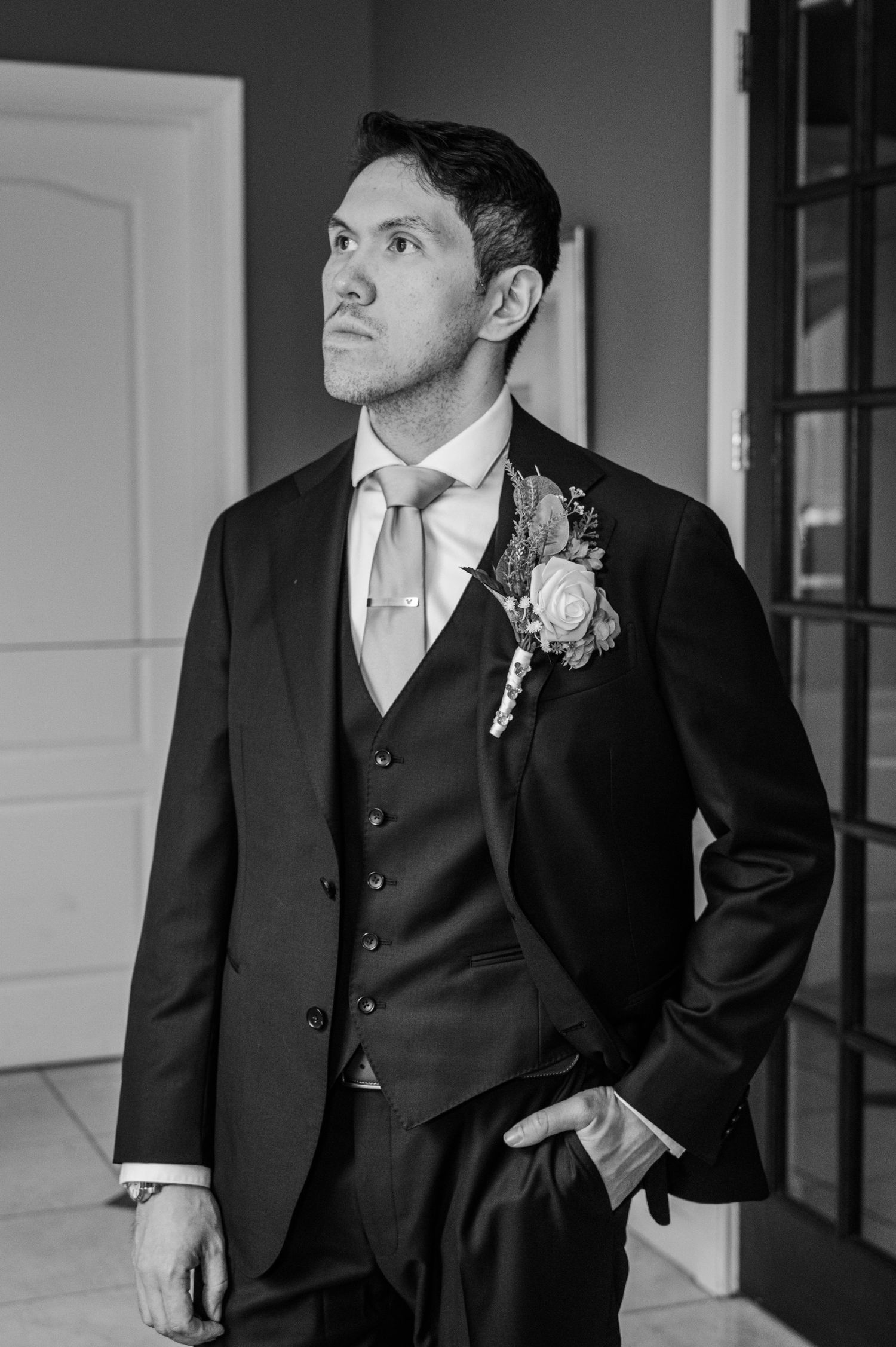 Elegant groom in a black three-piece suit with boutonniere and tie, posing in black and white wedding portrait.