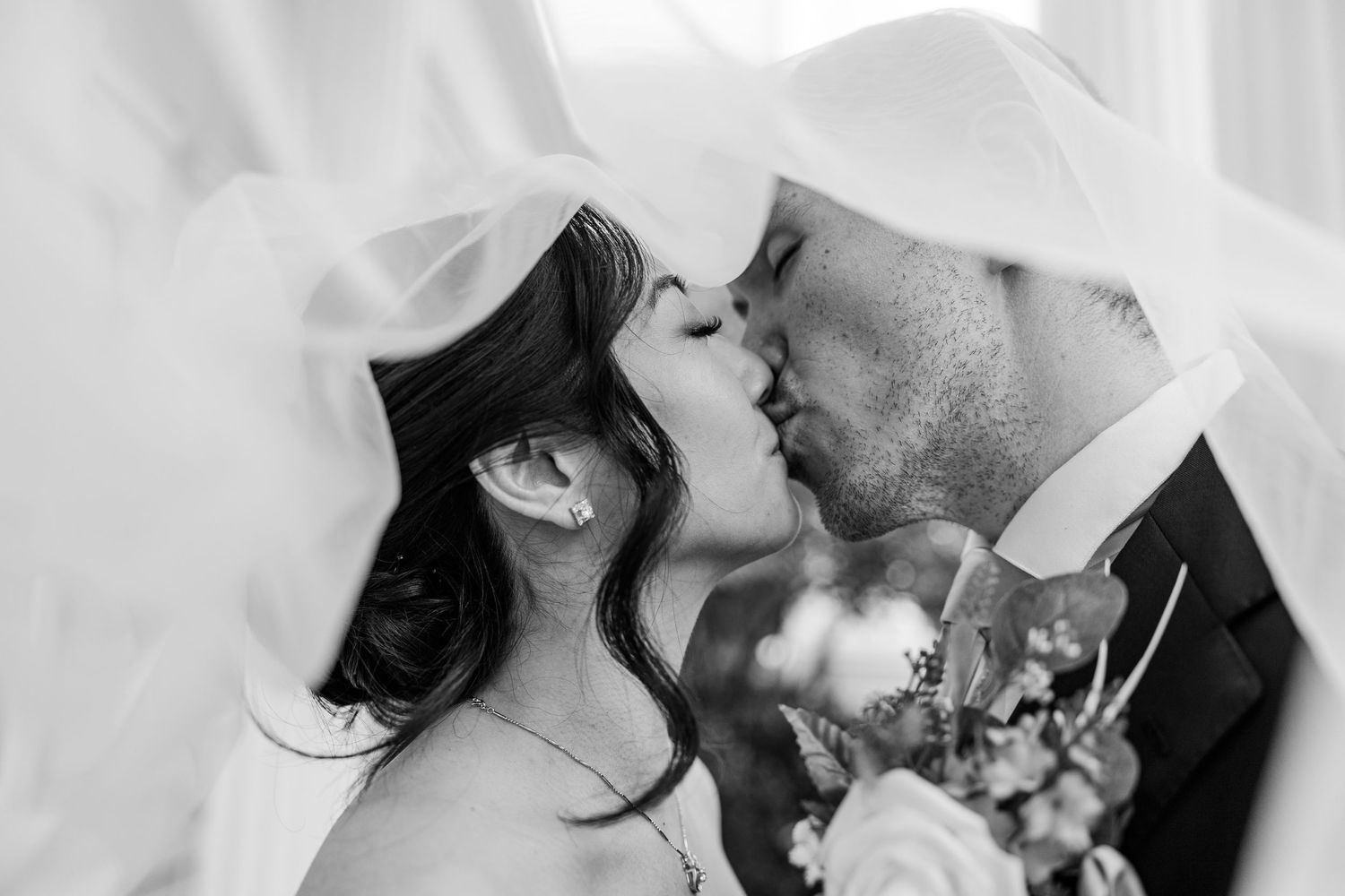 Black and white photo of a bride and groom sharing a romantic kiss under a flowing veil on their wedding day.