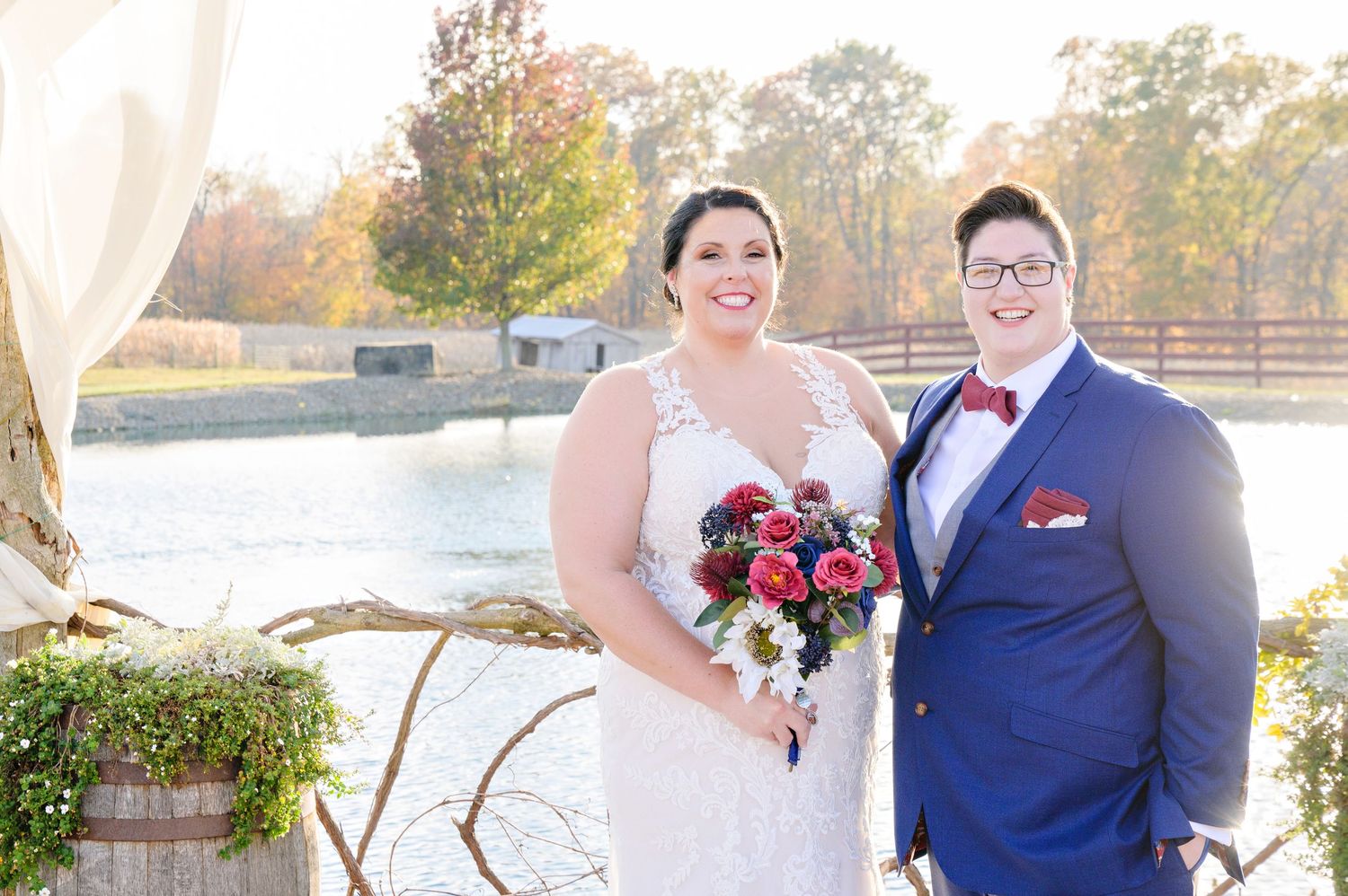 Two brides smiling and standing near the pond at Peacock Ridge after their wedding ceremony in the same spot in October.