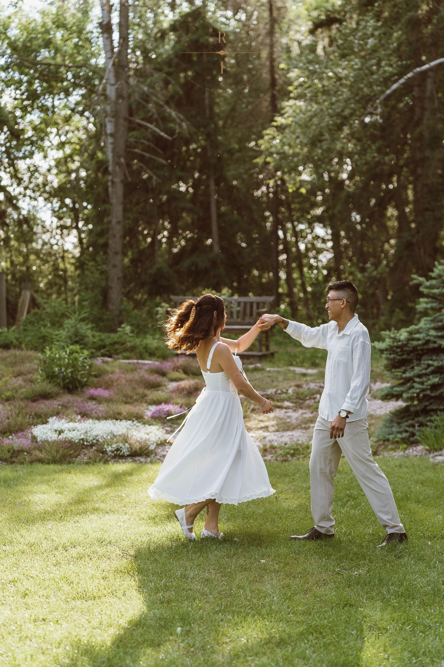 Couple dancing together on green lawn in garden with trees and purple flowers in background on sunny day.
