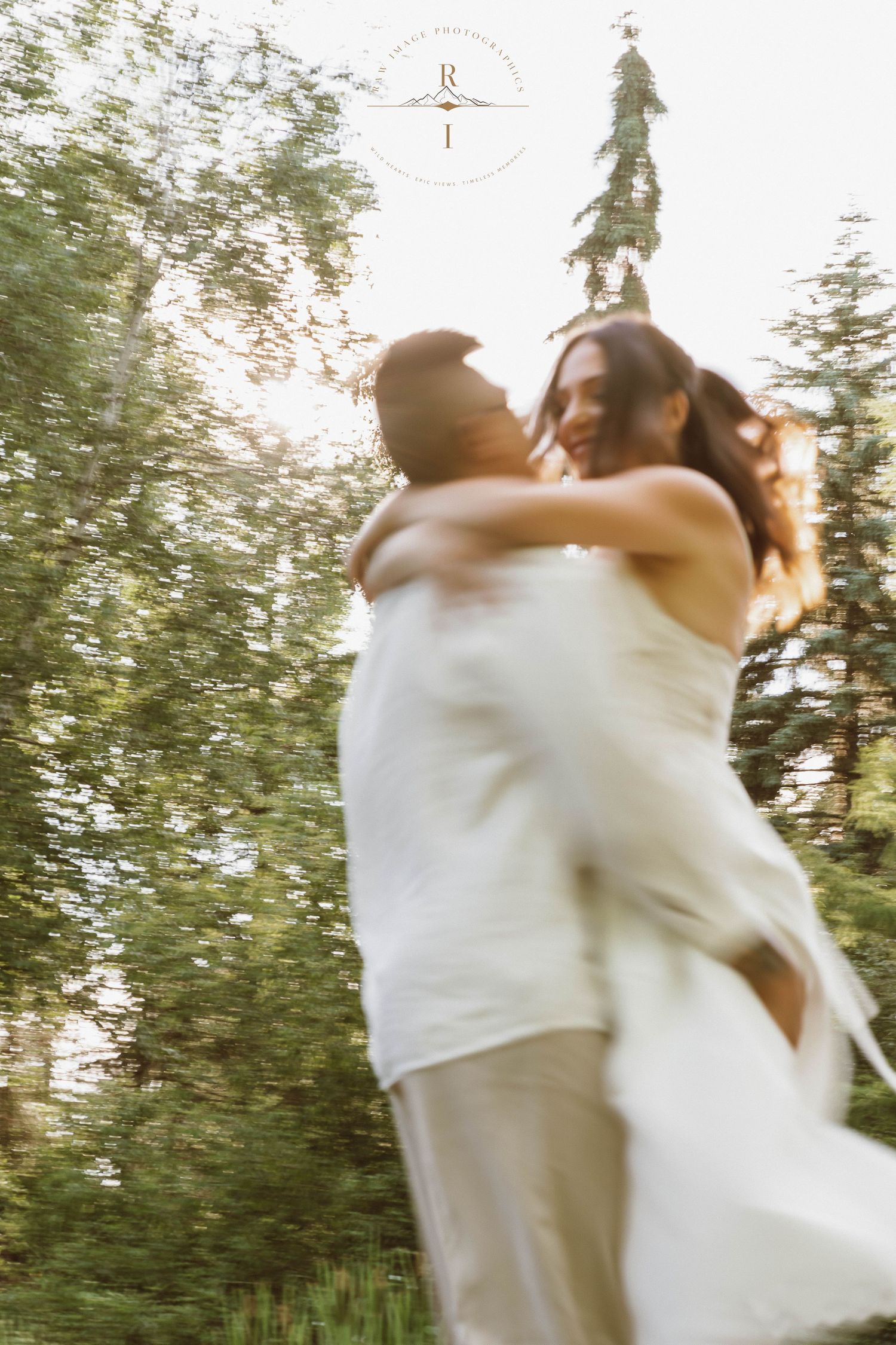 A joyful couple in white attire embraces and spins outdoors among lush green trees in a dreamy, motion-blurred wedding photo.