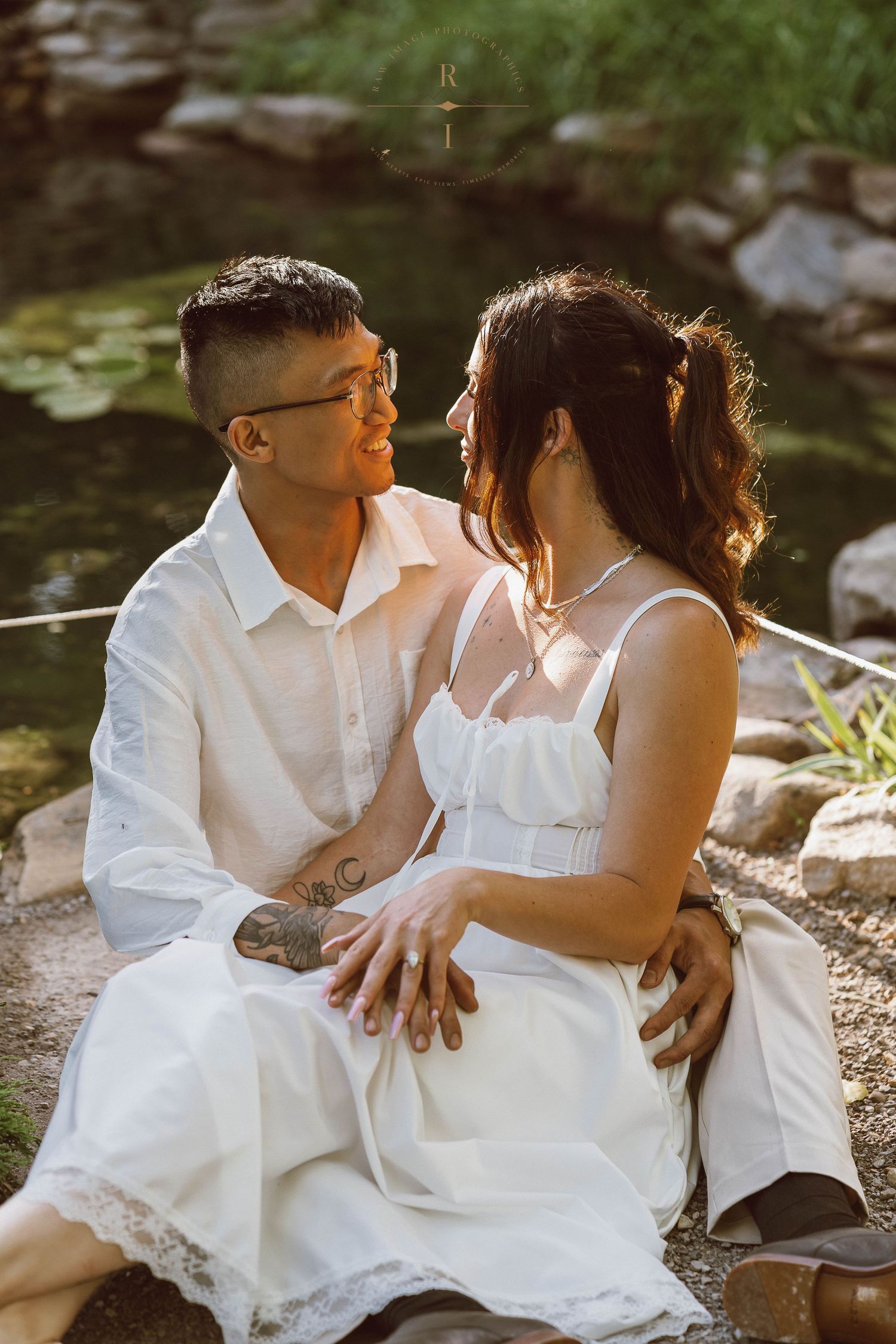 Couple in white outfits sitting together by a pond, sharing a romantic moment in a sunlit garden setting.