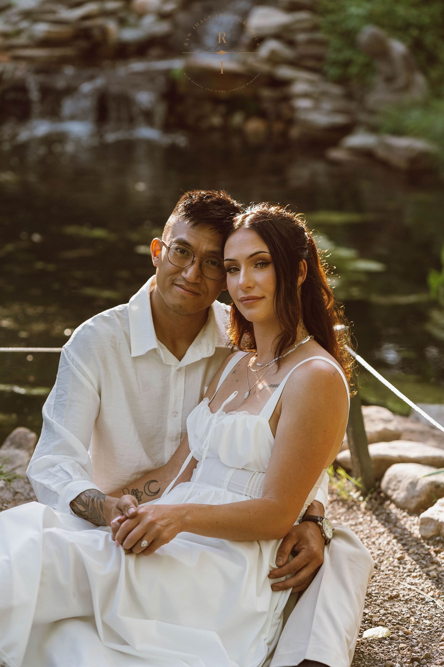 Couple in white attire sitting together near a waterfall, sharing a romantic moment outdoors.