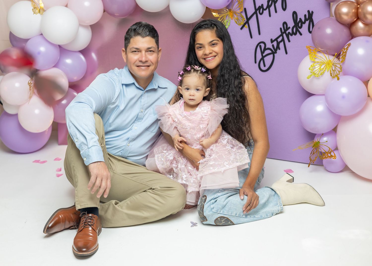 Family celebrating baby girl’s first birthday with balloons and Happy Birthday backdrop.