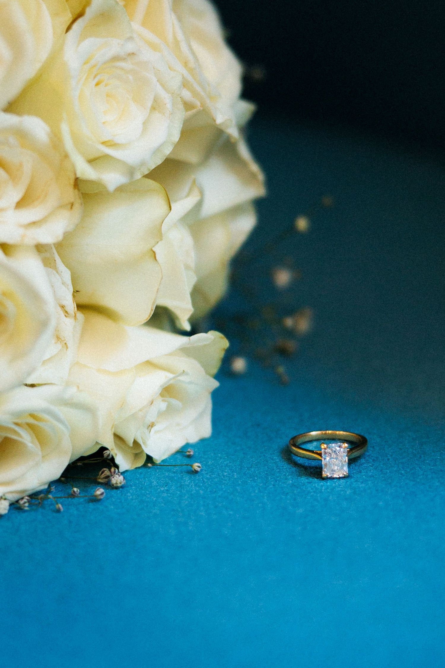 Gold engagement ring with diamond solitaire beside white rose bouquet on blue background.