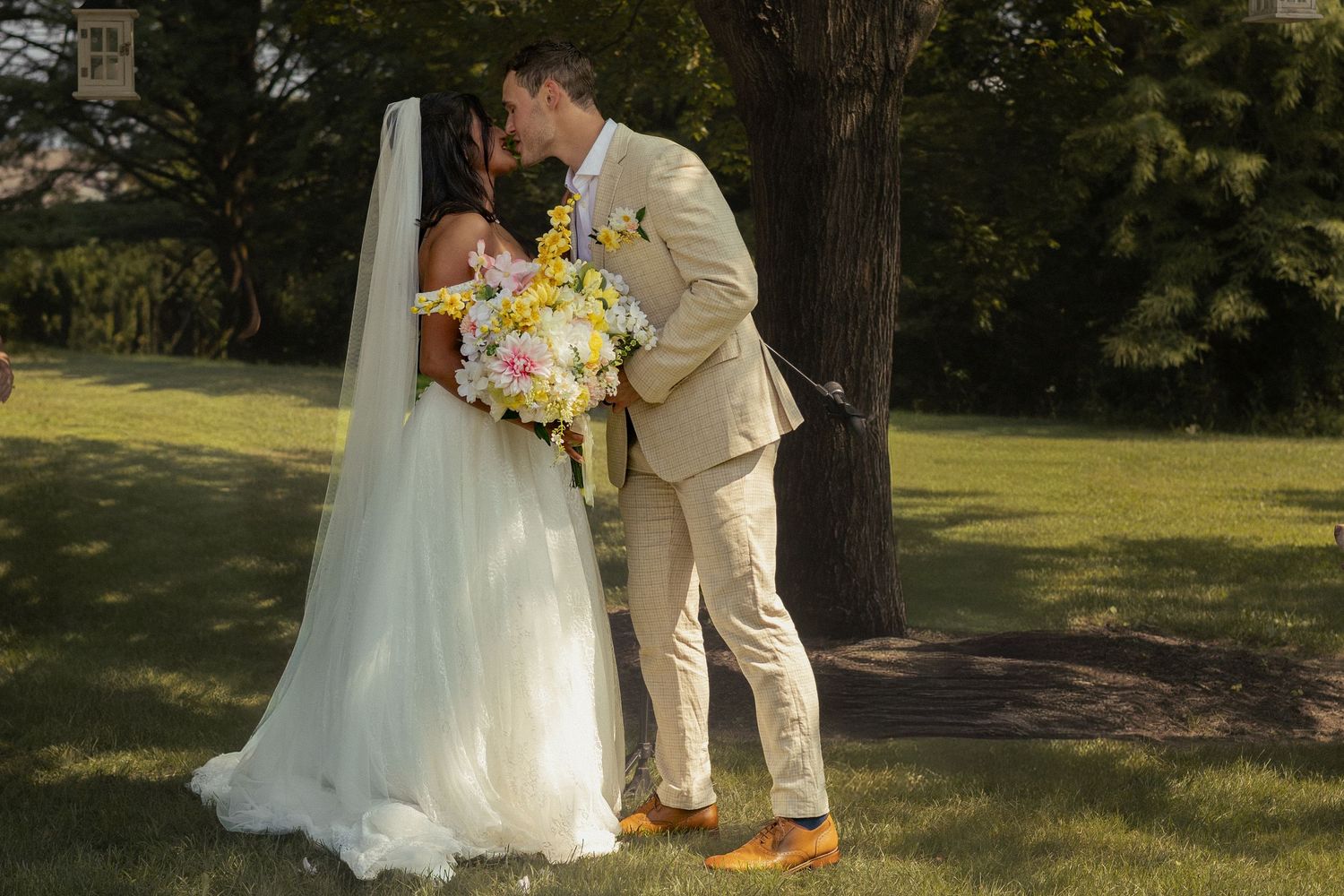 A bride and groom share their first kiss under a shady tree. The brides yellow and pastel tone bouquet is visible between them