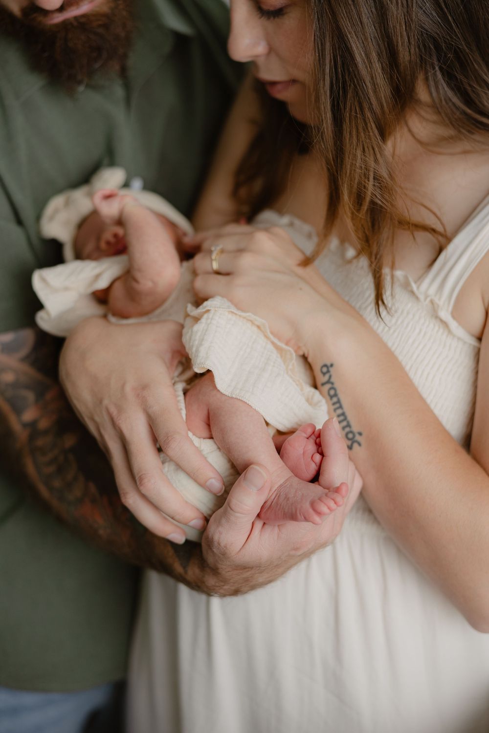 Parents tenderly cradling their newborn baby, mother wearing cream dress with tattoo visible, father in green shirt.