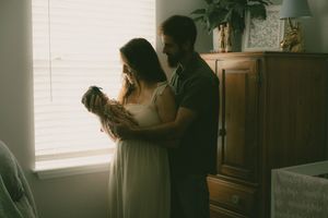 A couple stands by a window holding their newborn baby in a warmly lit bedroom with wooden furniture.