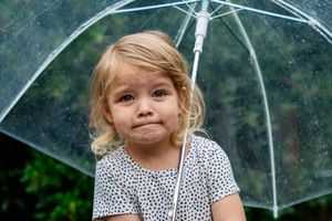 A cute toddler girl with blonde hair holds a clear umbrella in the rain, making an adorable pouty face outdoors.