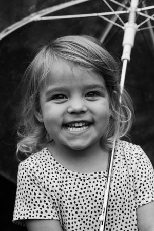 Happy toddler girl in polka dot dress holding a transparent umbrella, smiling in black and white portrait.