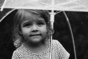 Black and white portrait of a young girl holding a transparent umbrella, looking up with a gentle smile.
