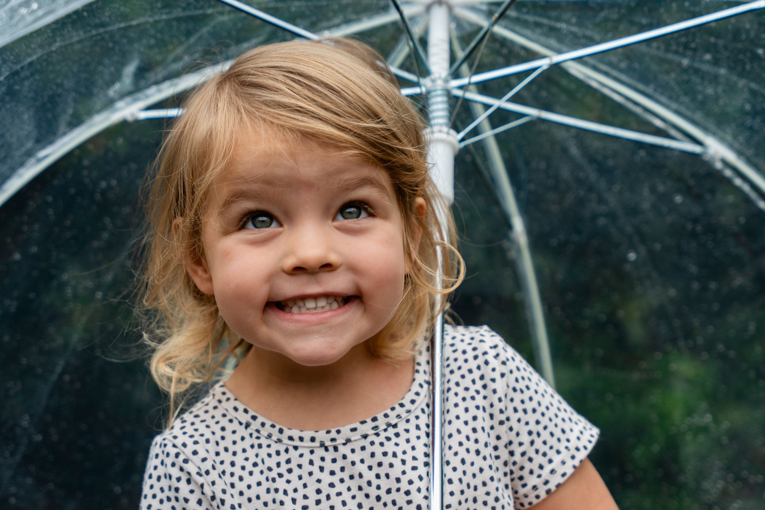 Smiling toddler girl with blonde hair holding a clear umbrella in the rain, wearing a white polka dot shirt.