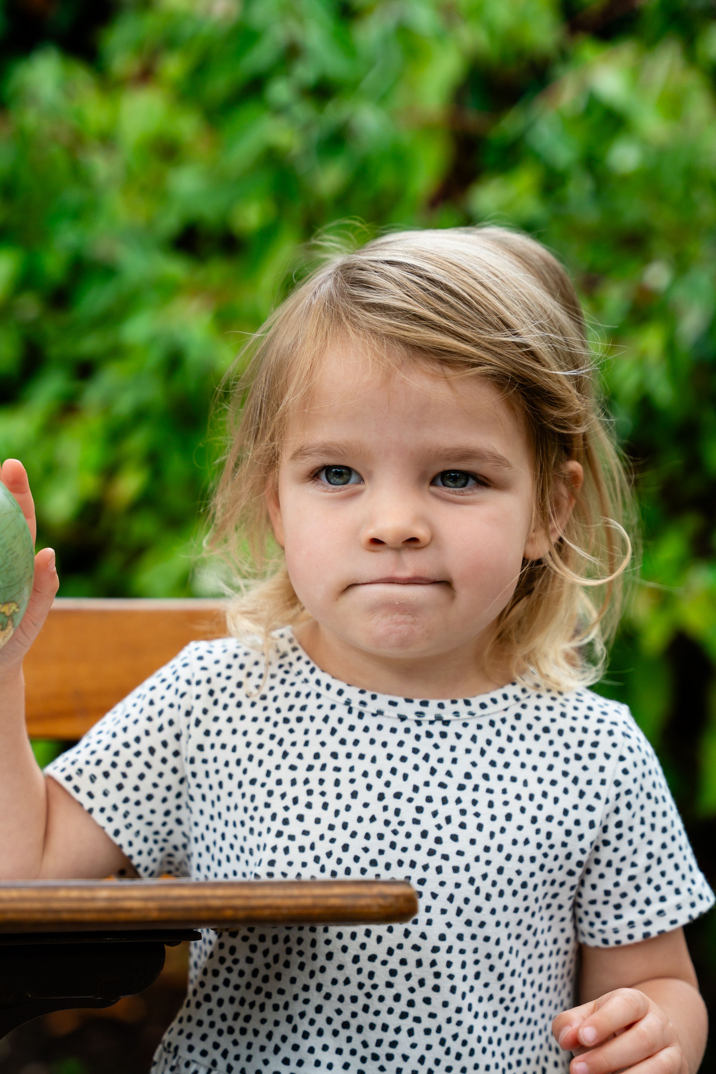 Young blonde toddler girl in polka dot dress sitting outdoors, holding a green ball with lush foliage background.