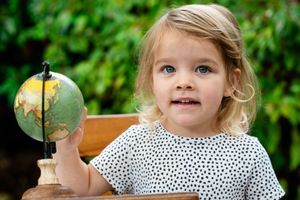 Blonde toddler girl holding a small globe outdoors, wearing a polka dot dress with green foliage background.