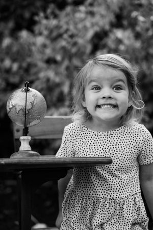 Smiling toddler girl in polka dot dress beside a small globe on a wooden desk, black and white photo.