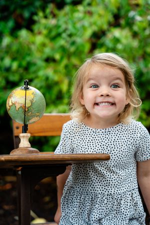 Smiling blonde toddler girl in polka dot dress sitting at wooden desk with small globe, outdoors.