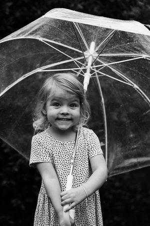 Smiling little girl in polka dot dress holding a clear umbrella in black and white photo.
