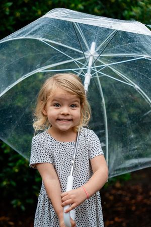 Smiling toddler girl with blonde curls holding a clear transparent umbrella outdoors in the rain.