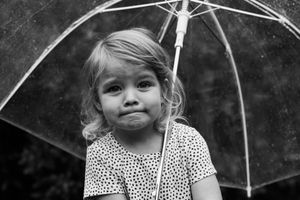 Young blonde girl holding a clear umbrella in the rain, wearing a polka dot dress, black and white photo.